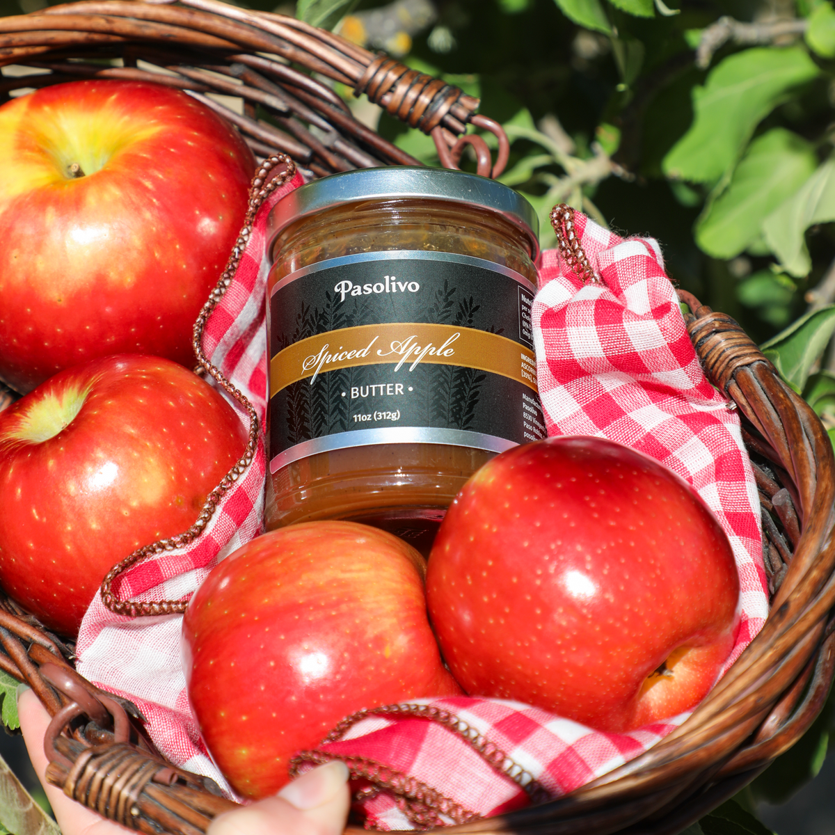 A jar of Pasolivo Spiced Apple Butter sits in a brown woven basket lined with red-and-white checkered cloth and surrounded by red apples, with green apple leaves in the background.