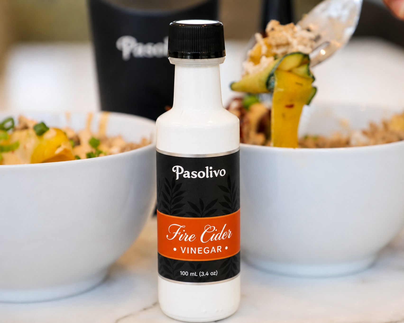 A bottle of Pasolivo Fire Cider Vinegar stands on a marble countertop between two white bowls of food, with a hand holding a spoon above one bowl.