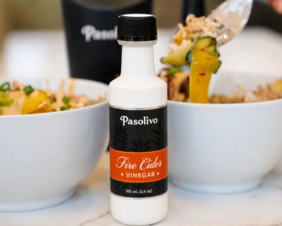 A bottle of Pasolivo Fire Cider Vinegar stands on a marble countertop between two white bowls of food, with a hand holding a spoon above one bowl.