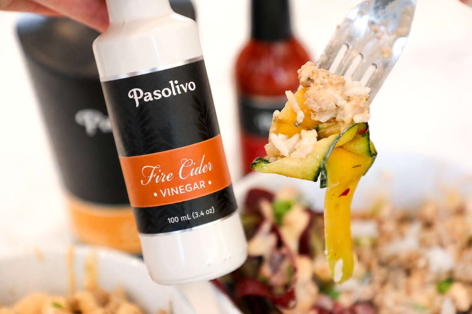 A hand holds a bottle of Pasolivo Fire Cider Vinegar in the foreground, with a fork lifting a piece of carrot topped with rice, turkey, and bang bang sauce over a bowl in a blurred kitchen setting.