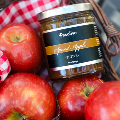 A jar of Pasolivo Spiced Apple Butter nestled in a wicker basket lined with a red and white checkered cloth and surrounded by fresh red apples.