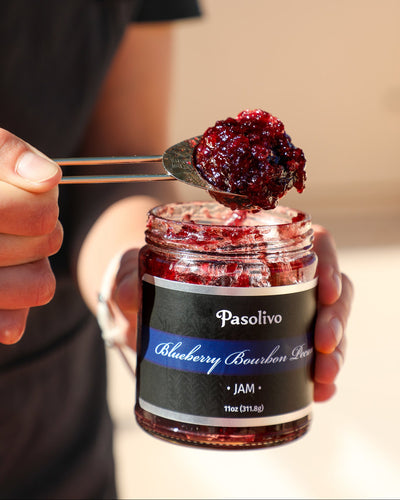 Person holding a jar of Pasolivo Blueberry Bourbon Pecan Jam with a spoonful of jam.