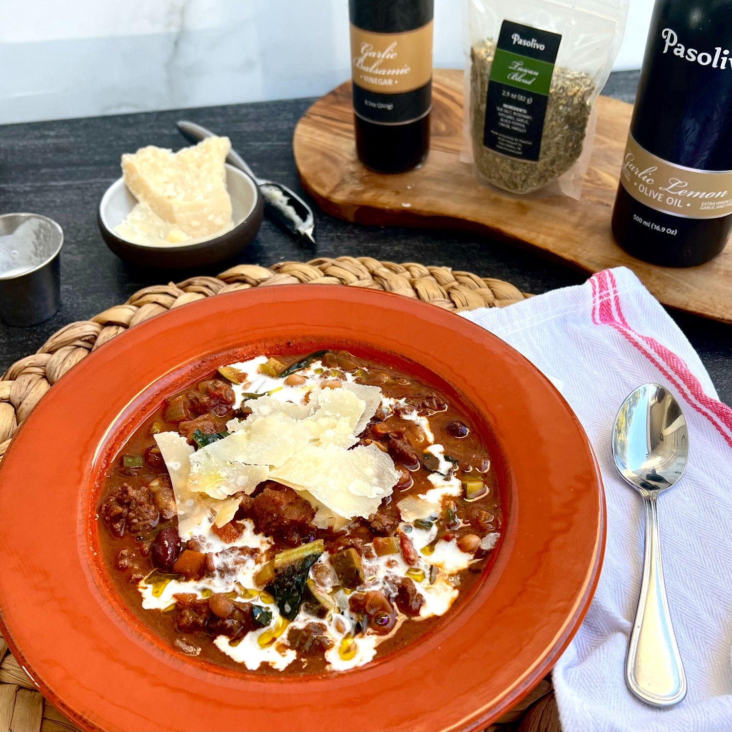 Pasolivo Tuscan Chili Recipe on a table next to a spoon, cheese with a cheese grater, Garlic Balsamic, Tuscan Blend, and Pasolivo Garlic Lemon Olive Oil