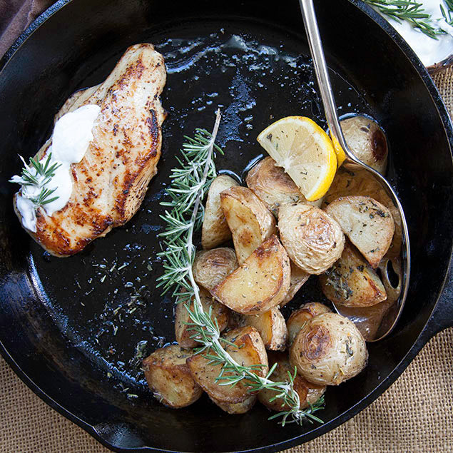 Pasolivo Yogurt & Citrus Olive Oil Chicken Recipe in a pan with potatoes and a serving spoon garnished with rosemary and a lemon wedge